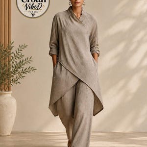 May include: A taupe-colored two-piece set consisting of a long-sleeved, wrap-style tunic with a high collar and side pockets, paired with matching wide-leg trousers. The outfit features a textured fabric and is worn with simple tan sandals.
