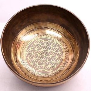 May include: A round, metallic singing bowl with a dark bronze exterior and a polished gold interior. The center of the bowl features an etched flower of life geometric pattern, surrounded by a decorative border of repeating petal motifs.
