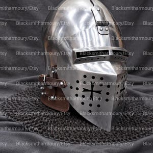 May include: A polished steel bascinet helmet featuring a hinged visor with a horizontal eye slit and a lower face plate with circular ventilation holes and a central cross emblem. Brown leather straps are attached to the side of the metal helmet.