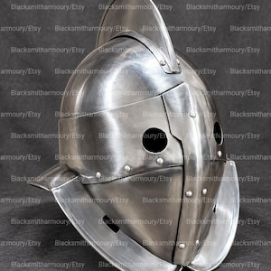 Könnte beinhalten: Ein polierter Morion-Helm aus Stahl mit einem hohen Mittelkamm, einer geschwungenen Krempe und einer Frontplatte mit zwei kreisförmigen Sehschlitzen. Die Metalloberfläche ist glatt und reflektierend, befestigt mit sichtbaren Nieten.