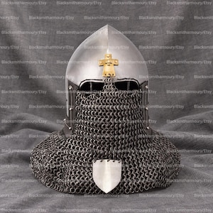 May include: A polished steel medieval-style helmet featuring a rounded dome, a gold-toned cross emblem at the forehead, and a protective chainmail aventail covering the face and neck, finished with a small shield-shaped metal plate at the center.