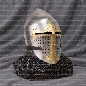 May include: A polished steel medieval-style helmet featuring a brass cross-shaped faceplate with decorative fleur-de-lis accents, a series of ventilation holes, a dark leather chin strap, and a black chainmail aventail covering the neck area.