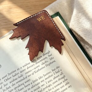May include: A dark brown leather corner bookmark shaped like a maple leaf with white stitching along the edges. The letters WE are embossed in gold on the top section of the bookmark, which is placed over the corner of an open book page.