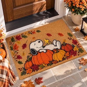 Op de afbeelding: Een rechthoekige kokosmat met een bedrukte illustratie van Snoopy en Woodstock die op oranje pompoenen liggen. Het ontwerp is omringd door een rand van kleurrijke herfstbladeren in rood, oranje en groen, tegen een natuurlijke beige achtergrond.