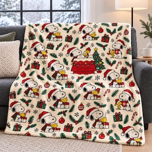 May include: A cream-colored fleece throw blanket featuring a repeating pattern of Snoopy and Woodstock in Santa hats, surrounded by Christmas trees, red ornaments, green gift boxes, candy canes, holly leaves, and white snowflakes.