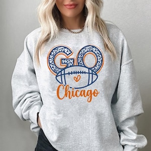 May include: A light gray crewneck sweatshirt featuring a graphic print on the chest. The design includes the word GO in blue leopard print, a blue football with an orange heart, and the word Chicago written in orange cursive script lettering.
