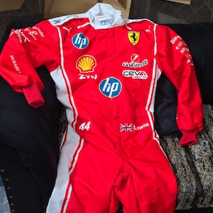 Peut inclure: Une combinaison de course rouge et blanche arborant divers logos de sponsors tels que HP, Shell, UniCredit et CEVA Logistics. La combinaison présente le numéro 44, un drapeau britannique, le nom Lewis et un emblème Ferrari sur le torse.