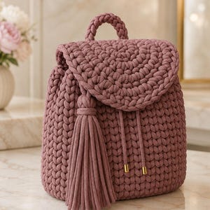 May include: A dusty rose crochet backpack featuring a thick braided top handle, a rounded flap closure, and a long matching tassel attached to the side. The bag is constructed from chunky yarn with a visible knit texture and includes two drawstring cords.