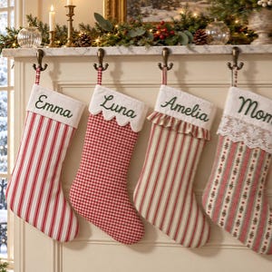Puede incluir: Cuatro calcetines de Navidad colgados de ganchos de metal. El primero es de rayas rojas y blancas con 'Emma' bordado. El segundo es de cuadros vichy rojos y blancos con 'Luna' bordado. El tercero es de rayas con un volante y 'Amelia' bordado. El cuarto tiene encaje y 'Mom' bordado.