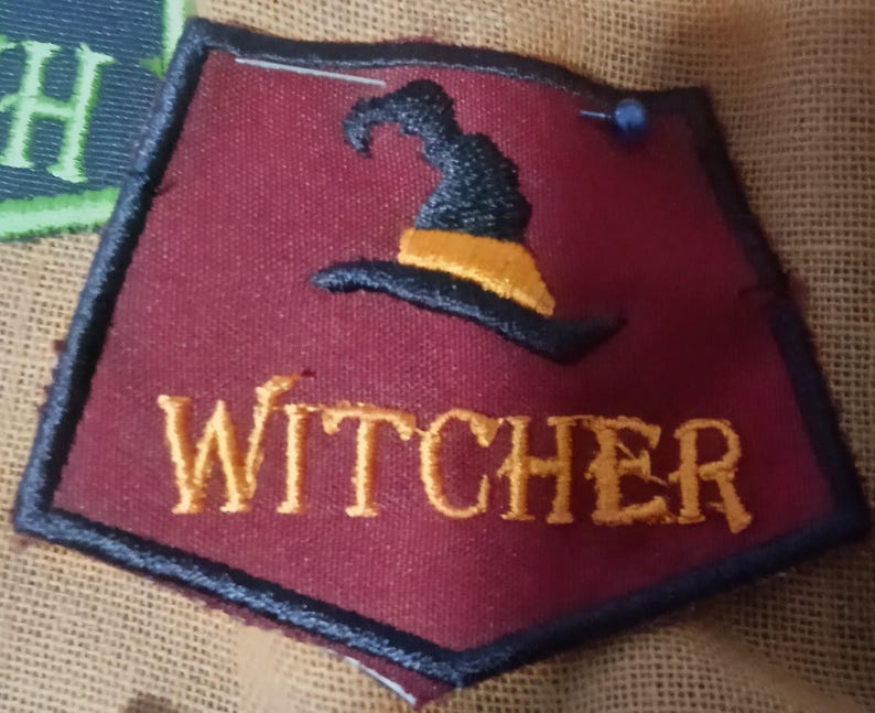 Pode incluir: Um emblema de tecido em forma de escudo em vermelho escuro com uma borda preta. Apresenta um chap&eacute;u de bruxa preto bordado com uma faixa laranja acima do texto amarelo bordado WITCHER. Um pequeno alfinete azul est&aacute; preso no canto superior.