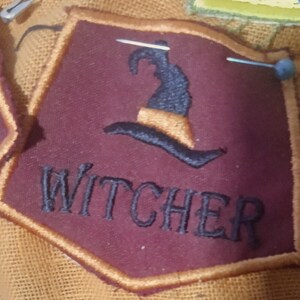 Pode incluir: Um emblema em forma de escudo na cor bord&ocirc; escuro com uma borda dourada. O emblema apresenta um &iacute;cone de chap&eacute;u de bruxa bordado em preto acima da palavra WITCHER em letras mai&uacute;sculas pretas. Dois alfinetes de costura prateados est&atilde;o fixados.