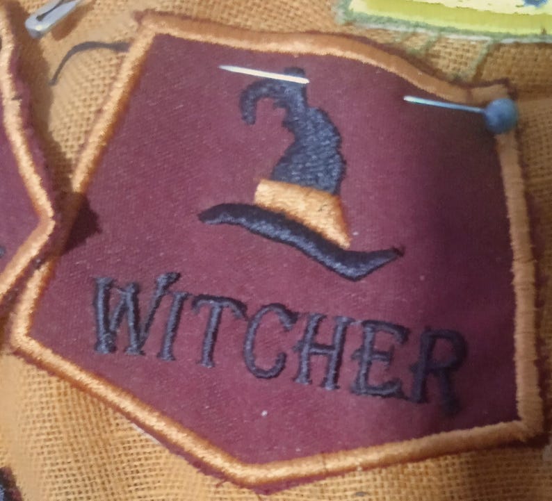 Pode incluir: Um emblema em forma de escudo na cor bord&ocirc; escuro com uma borda dourada. O emblema apresenta um &iacute;cone de chap&eacute;u de bruxa bordado em preto acima da palavra WITCHER em letras mai&uacute;sculas pretas. Dois alfinetes de costura prateados est&atilde;o fixados.
