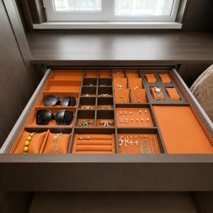 May include: A dark wood jewelry drawer organizer featuring orange velvet-lined compartments. It contains two pairs of black sunglasses, gold necklaces, earrings, bracelets, rings, and a wristwatch displayed on orange cushions and in divided sections.