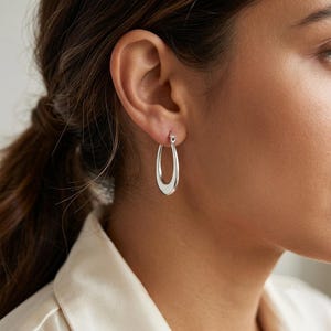 May include: A polished silver-toned hoop earring with a smooth, elongated teardrop shape. The earring features a hinged closure mechanism at the top, securing it through the earlobe. The metal surface is reflective and solid with a curved design.