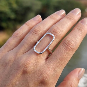 Op de afbeelding: Een zilveren ring met een groot, open, verticaal georiënteerd rechthoekig frame. De band is dun en gepolijst en sluit aan op de basis van de langwerpige rechthoekige vorm, die met een gladde, metalen afwerking op de vinger rust.