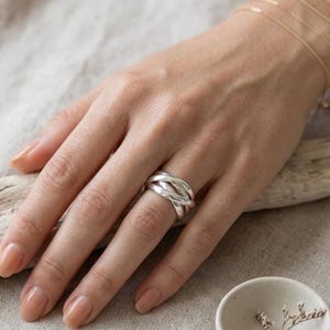 Può includere: Un anello in argento lucido caratterizzato da un design a fasce multiple con diversi anelli intrecciati e sovrapposti che creano un aspetto ampio e stratificato. Il metallo presenta una finitura liscia e riflettente che cattura la luce.