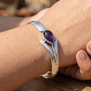 Puede incluir: Una pulsera de brazalete de plata con una banda lisa y pulida que se divide en dos brazos. Una piedra preciosa de amatista morada en forma de lágrima está engastada en el centro, acentuada por un detalle de cuerda de plata trenzada.