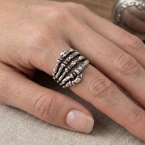 May include: A silver-toned ring featuring a multi-band design that resembles a skeletal hand wrapping around the finger. The metal has an oxidized finish to highlight the detailed bone structures, including small skull motifs and articulated joints.