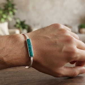 May include: A silver cuff bracelet featuring a rectangular, horizontally oriented turquoise stone inlay with dark veining. The metal band is smooth and polished, encircling the wrist with a clean, open-ended design that highlights the stone.