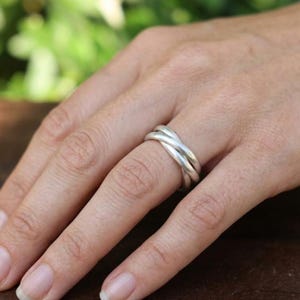 Peut inclure: Une bague en argent au design triple anneau composé de trois bandes métalliques lisses et polies qui s'entrelacent pour former un seul bijou cohérent porté au doigt, présentant une finition métallique brillante et réfléchissante.