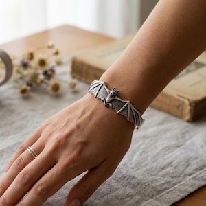 May include: A silver-toned metal cuff bracelet featuring a detailed bat design with outstretched wings. The bat is positioned in the center of the cuff, which wraps around the wrist. A thin, simple silver-toned ring is worn on the ring finger.