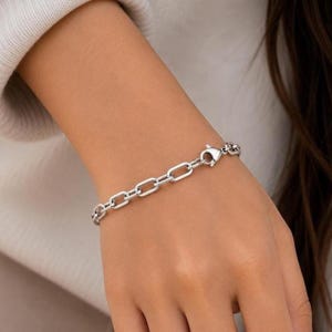 May include: A silver-toned chain bracelet featuring elongated, rectangular links with rounded edges. The bracelet is secured with a lobster claw clasp, which is clearly visible as part of the chain design, creating a uniform and metallic appearance.