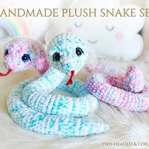 May include: A handmade plush snake set featuring two crocheted snakes with soft, variegated yarn in shades of pink, blue, and white. One snake has two heads with dark eyes and red felt tongues, while the other is coiled. Text reads HANDMADE PLUSH SNAKE SET.