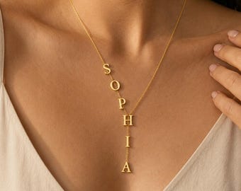 Puede incluir: Un collar de tono dorado con una cadena fina que presenta el nombre SOPHIA escrito en letras mayúsculas verticales con serifa. Las letras cuelgan directamente de la cadena, creando un diseño lineal que cae verticalmente sobre el pecho.