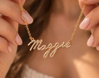 Op de afbeelding: Een goudkleurige ketting met een naamplaatje in handschriftstijl dat de naam Maggie spelt. De hanger is over het gehele oppervlak van de letters ingelegd met kleine, heldere, fonkelende steentjes en hangt aan een fijne, delicate ketting.