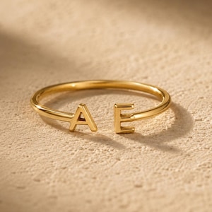 May include: A gold-toned open ring featuring two capital letters, A and E, positioned at the ends of the band. The ring has a smooth, polished finish and a simple, circular design that remains open between the two letters.