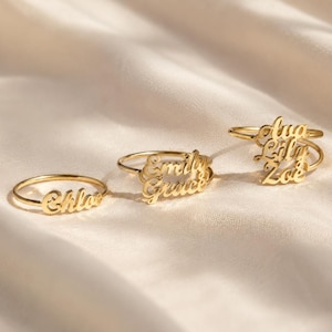 Peut inclure: Trois bagues dorées avec des inscriptions en lettres cursives personnalisées. La première bague porte le prénom Chloe, la deuxième présente les prénoms Emily et Grace superposés, et la troisième affiche les prénoms Ava, Lily et Zoe.
