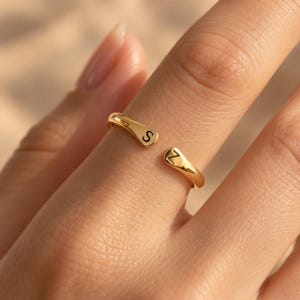 May include: A gold-toned open ring featuring two tapered ends, each engraved with a single black letter. One end displays the letter S, while the other end displays the letter Z. The band has a smooth, polished finish and a simple, adjustable design.