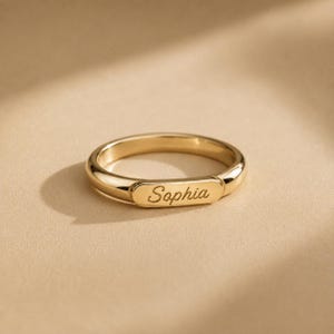 May include: A polished gold-tone ring featuring a smooth, rounded band and a flat, rectangular center plate. The name Sophia is engraved in a cursive script font across the center of the plate, creating a personalized piece of jewelry.