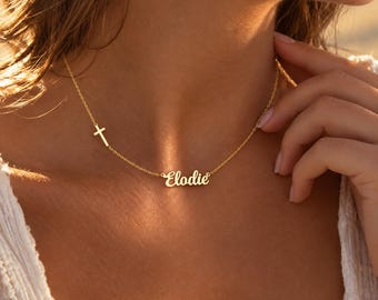 Puede incluir: Un collar de tono dorado que presenta una cadena delicada con un pequeño dije de cruz posicionado en el lado izquierdo y un colgante central con el nombre Elodie en una fuente de escritura cursiva, descansando sobre el cuello.
