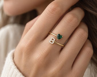 Op de afbeelding: Een open gouden ring met een letter B ingelegd met kleine heldere steentjes aan het ene uiteinde en een enkele donkergroene hartvormige steen aan het andere uiteinde. De ringband is dun en gepolijst voor een verfijnde uitstraling.