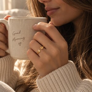 Woman wearing gold double initial ring while holding a mug that reads Good Morning