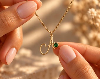 Op de afbeelding: Een goudkleurige ketting met een hanger in de vorm van een cursieve letter A. De hanger is versierd met een kleine, ronde, groenkleurige edelsteen in een zetting. De ketting hangt aan een fijne goudkleurige boxketting.