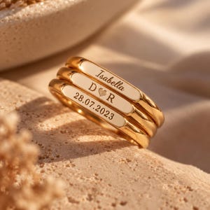 May include: A stack of three gold-toned rings with flat, rectangular faces. The top ring is engraved with the name Isabella, the middle ring features the letters D and R separated by a heart symbol, and the bottom ring displays the date 28.07.2023.
