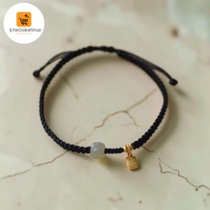 May include: A black braided cord bracelet featuring a single smooth, pale grey stone bead and a small, rectangular gold-toned metal charm with embossed characters. The bracelet has adjustable pull-string ends for a secure and flexible fit.