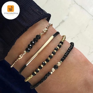 May include: A set of four bracelets featuring black faceted beads, gold-toned metallic beads, and a smooth gold-toned bar element. The bracelets are arranged in a stack, combining delicate gold-toned chains with black cord and small dark beads.