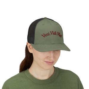 May include: An olive green trucker cap with a black mesh back and a stiff peak. The front panel features the embroidered text Veni, Vidi, Vici in a dark red, serif font. The cap has a structured crown and a matching olive green button on top.