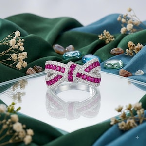 May include: A silver-toned ring shaped like a bow, featuring rows of vibrant pink circular gemstones bordered by small, clear white stones. The ring is positioned on a reflective surface, showcasing its intricate design and sparkling finish.