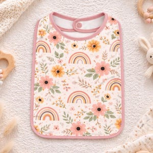 Può includere: Una bavaglina con sfondo bianco e un motivo ripetuto di fiori rosa e gialli, foglie verdi e piccoli arcobaleni. La bavaglina presenta un bordo rosa a tinta unita lungo i contorni e una chiusura con bottone a pressione sul collo.