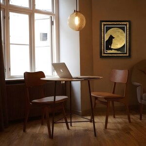 May include: A framed wall art piece featuring a black silhouette of a wolf sitting on a rocky outcrop against a large, textured gold full moon. The artwork is enclosed in a black frame with gold inner detailing, displayed on a wall.