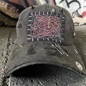 Puede incluir: Una gorra de béisbol gris carbón desgastada con un parche rectangular de tela bordado con hilos rojos y blancos. La gorra está decorada con púas metálicas plateadas, varios imperdibles y ojales metálicos a lo largo de la visera y la corona.