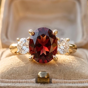Puede incluir: Un anillo de oro con una gran piedra preciosa granate de color rojo intenso y corte ovalado en el centro, flanqueada por dos diamantes blancos transparentes de corte redondo más pequeños. El anillo se presenta en un joyero de terciopelo beige.