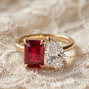 Puede incluir: Un anillo de oro con dos piedras prominentes: un rubí rojo intenso de corte esmeralda rectangular y un diamante blanco transparente de corte en forma de pera. Las piedras están engastadas una al lado de la otra en una banda de oro pulido.