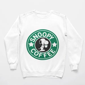 Puede incluir: Una sudadera blanca de manga larga con un gráfico circular verde y blanco en el pecho. El gráfico muestra un perro de dibujos animados sosteniendo una taza, rodeado por el texto SNOOPY COFFEE en un borde circular con dos pequeñas estrellas blancas.