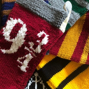 Puede incluir: Un conjunto de cinco calcetines tejidos con diferentes colores y símbolos de las casas de Hogwarts. Los calcetines son rojos, amarillos, negros, verdes y grises con detalles blancos. El calcetín rojo tiene el número "9 3/4" tejido en él.