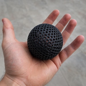 Pode incluir: Um objeto esférico preto com uma estrutura de treliça texturizada semelhante a um favo de mel. A superfície da esfera é composta por um padrão repetitivo de pequenas aberturas hexagonais interconectadas, criando um aspecto poroso.